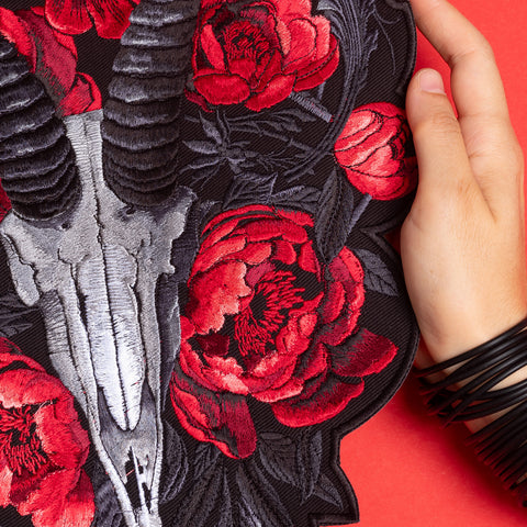 A hand holds the Blesbok Skull and Flowers Embroidered Patch, showcasing a silver ram skull with black horns and vivid red flowers—an ideal accessory for goth fashion fans.