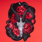 The Blesbok Skull and Flowers Embroidered Patch features a blesbok skull with curved horns, red peonies, and dark foliage on black—perfect for goth fashion and shown on a bold red background.