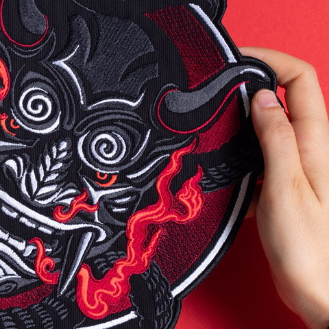 A hand holds the Oni Japanese Demon Embroidered Patch, inspired by Asian folklore, with swirling white and red accents, sharp teeth, and red flames on a vivid red background.