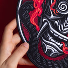 A hand holds the Oni Japanese Demon Embroidered Patch, inspired by Asian folklore, showcasing a stylized fierce face with sharp teeth, swirling patterns, and vivid red accents on a black and white background.