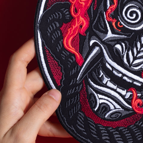 A hand holds the Oni Japanese Demon Embroidered Patch, inspired by Asian folklore, showcasing a stylized fierce face with sharp teeth, swirling patterns, and vivid red accents on a black and white background.