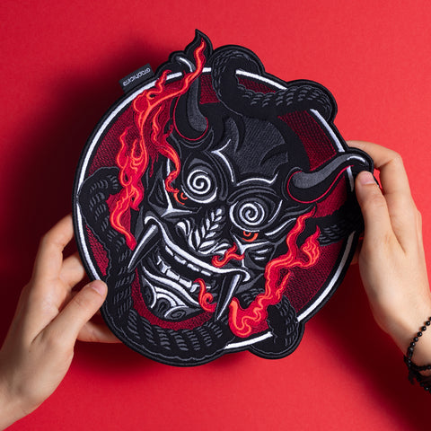 Two hands hold the Oni Japanese Demon Embroidered Patch, which features a stylized black demonic mask with swirling eyes, fiery red accents, horns, and a black snake, inspired by Asian folklore, set against a bold red background.