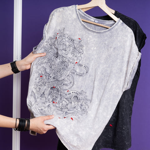 A person holds up the Japanese Koi Fish Embroidered Female T-shirt, a gray 100% cotton short-sleeve with black dragon and wave design, text ribbons, and red accents. A black T-shirt hangs behind it, set against a purple background.