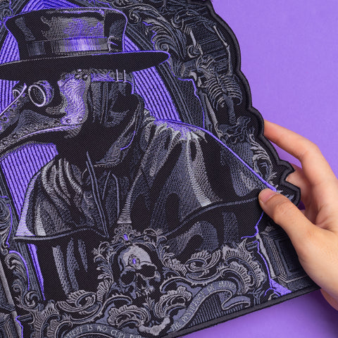 A hand holds the Plague Doctor Embroidered Back Patch, featuring a beaked mask, wide-brimmed hat, ornate designs, and a skull—an ideal Victorian cosplay accessory—set against a vivid purple background.