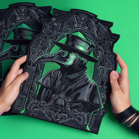 Two hands hold ornate black and gray Plague Doctor Embroidered Back Patches, outlined in neon green. Featuring a beaked mask and wide-brimmed hat, they make a striking Victorian cosplay accessory on a vibrant green background.