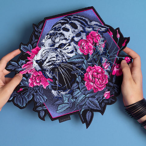 Two hands hold the Tiger and Roses Embroidered Patch, featuring a vibrant tiger among pink roses and green leaves on a blue background, showcasing vivid colors and detailed stitching for true Bohemian flair.