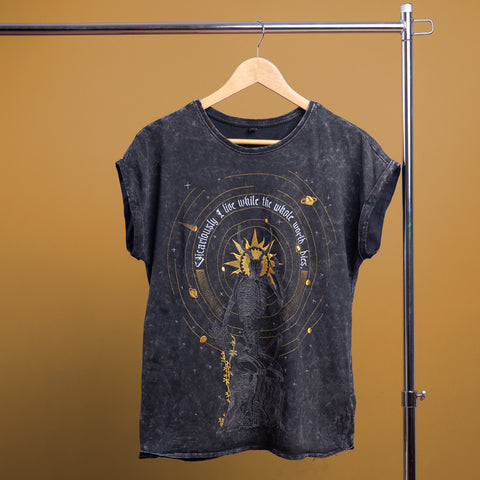 The Vicarious Embroidered Female T-shirt features a gold celestial design and the phrase “Living a life while the whole world dies” on black fabric, perfect for gothic fashion lovers. Shown hanging on a wooden hanger against a brown background.