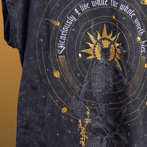 A close-up of the Vicarious Embroidered Female T-shirt, styled for gothic fashion, featuring an intricate gold and white skeleton with a scythe amid celestial motifs and the text “Vicariously I live while the whole world dies.”.