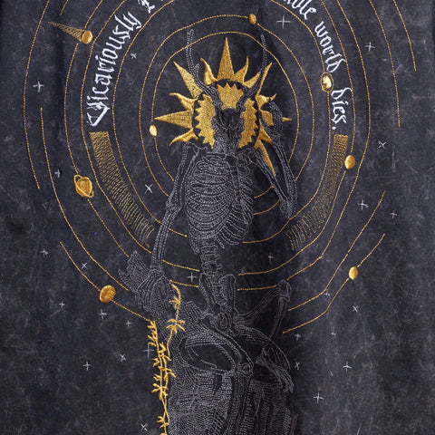 The Vicarious Embroidered Female T-shirt features a skeleton with a radiant sun halo, concentric yellow circles, and stars on a dark background. “Vicariously I live while the whole world dies” arcs above—a bold statement of gothic style.