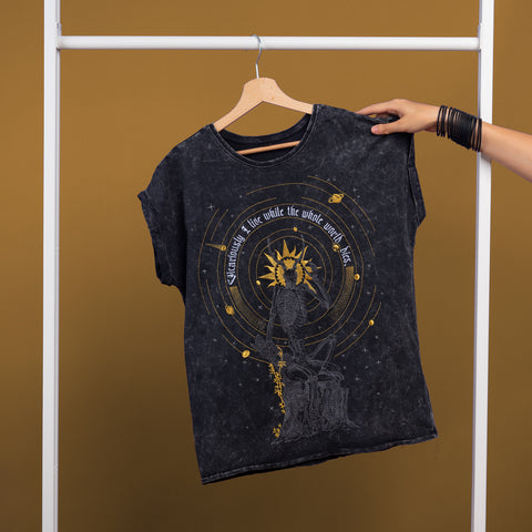 A hand holds the Vicarious Embroidered Female T-shirt, black and hanging against a brown background. Inspired by gothic fashion, it features gold sun and celestial embroidery with the phrase "As above, so below, the whole universe fits.