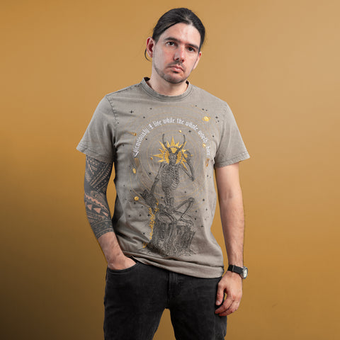 A man with long dark hair and a tattoo sleeve stands against a brown background, wearing the Vicarious Embroidered Unisex T-shirt, which features Victorian-style embroidery of a giraffe and white text. His style hints at subtle gothic fashion.