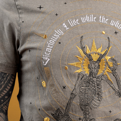 The Vicarious Embroidered Unisex T-shirt features a Victorian skeletal figure, golden sunburst, stars, and "Vicariously I live while the whole world dies" text. A tattooed arm adds dark art flair to this unique close-up.