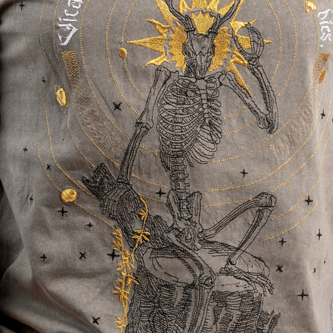 The Vicarious Embroidered Unisex T-shirt features a skeletal figure with animal horns on a rock, surrounded by golden circles and stars—a bold fusion of dark art and gothic fashion for a standout look.