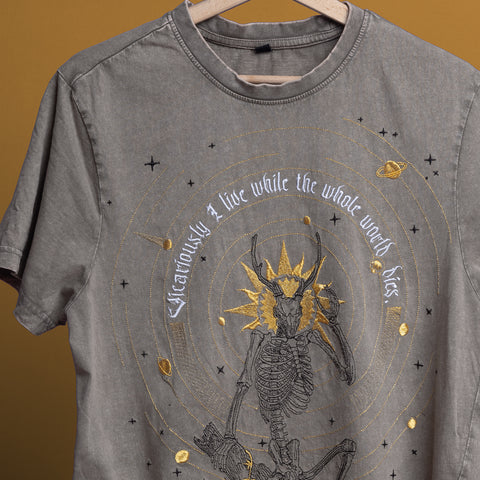 The Vicarious Embroidered Unisex T-shirt, shown in gray on a wooden hanger, features a dark art design of a skeletal figure with a golden crown, celestial symbols, and the text, "Vicariously, I live while the whole world dies.